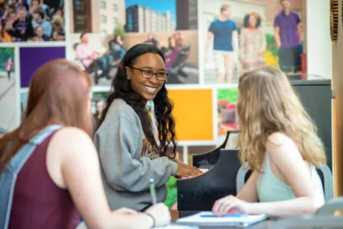 A smiling woman wearing glasses plays the piano while two other women, possibly discussing West Chester University housing, sit nearby talking and taking notes. They are indoors, with colorful photos of people on the wall behind them.