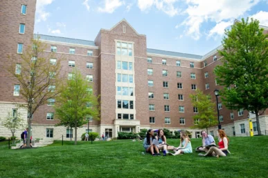A group of students sits on the grass chatting in front of a large West Chester University housing building with several windows on a sunny day, trees and a blue sky in the background.