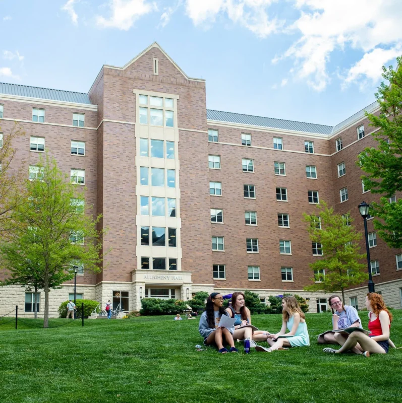 A group of students sits on the grass chatting in front of a large West Chester University housing building with several windows on a sunny day, trees and a blue sky in the background.