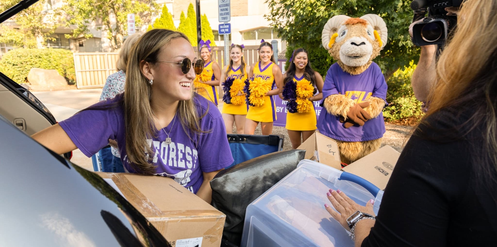 A smiling student wearing sunglasses unloads boxes from a car, assisted by another person, during West Chester University housing move-in. Cheerleaders and a lion mascot in purple and gold cheer nearby, with trees and a building in the background.