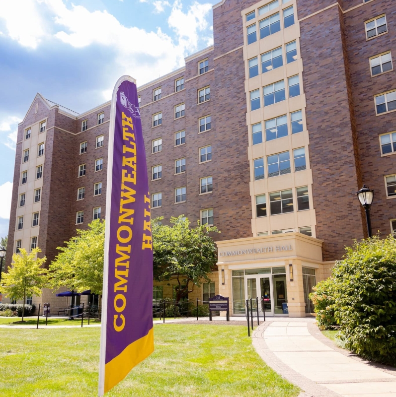 A purple and yellow flag reading COMMONWEALTH HALL stands on a grassy lawn in front of a large, multi-story brick building—one of the West Chester University housing options—with many windows and a sign over the entrance that also says Commonwealth Hall.