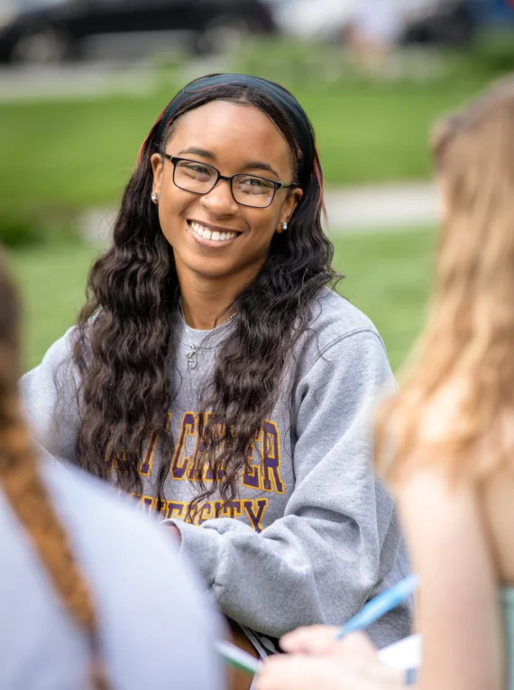 A young woman with long wavy hair and glasses smiles while sitting outdoors with others. Wearing a gray West Chester University housing sweatshirt, she holds a notebook, surrounded by green grass in the background.