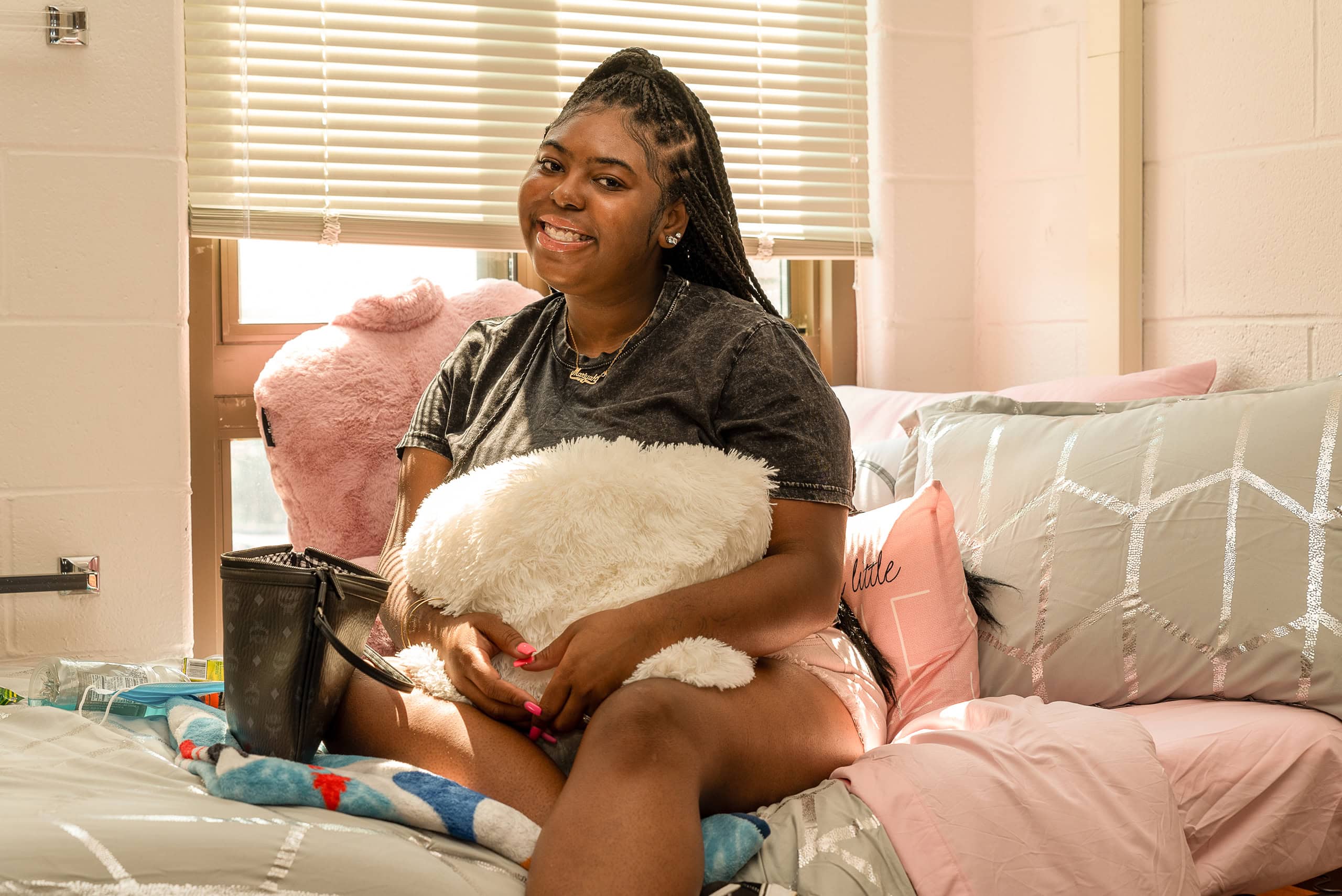 A young woman sits on a neatly made bed in a bright West Chester University housing dorm room, smiling and hugging a fluffy white pillow. Light streams through the blinds, illuminating the cozy, pastel-colored bedding and decor.