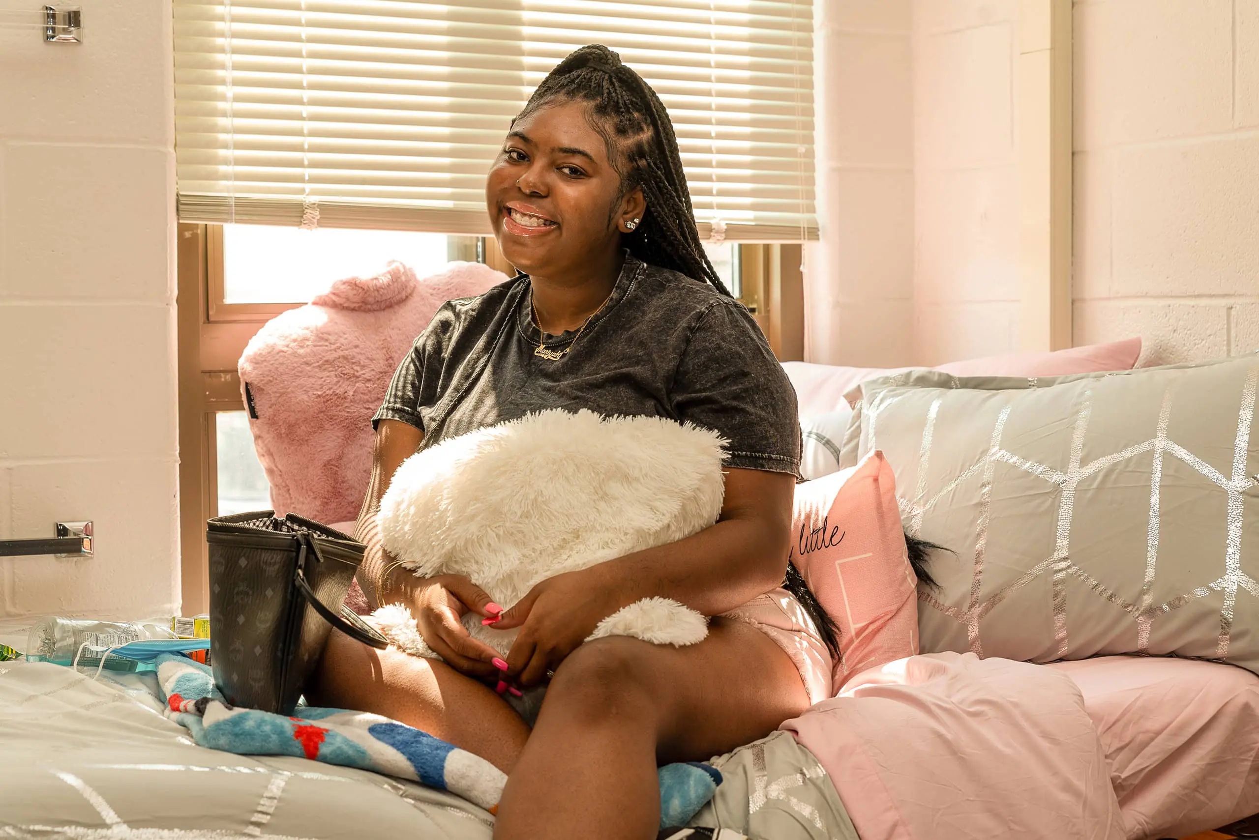 A young woman sits on a neatly made bed in a bright West Chester University housing dorm room, smiling and hugging a fluffy white pillow. Light streams through the blinds, illuminating the cozy, pastel-colored bedding and decor.