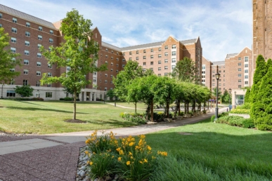 A landscaped courtyard with green lawns, yellow flowers, trees, and a pergola sits in front of large brick West Chester University housing buildings under a partly cloudy sky.