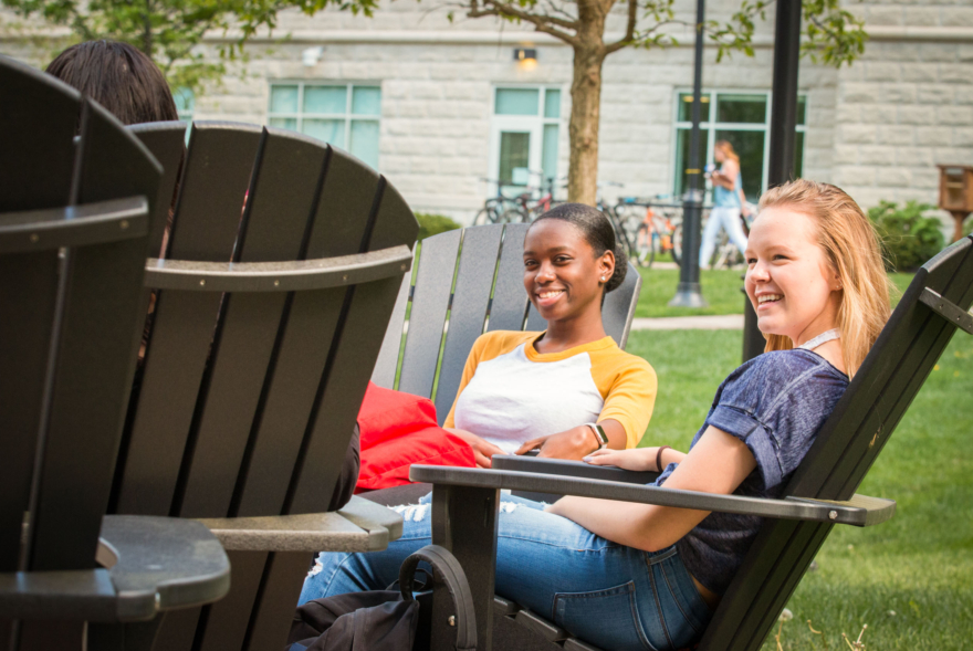 Two young women sit in Adirondack chairs on a sunny lawn, smiling and relaxing outside a building with large windows and a tree—capturing the welcoming atmosphere of West Chester University housing.