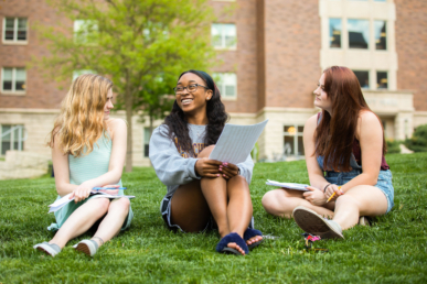 Three young women sit on grass outside a building, holding papers and talking. They appear to be studying, perhaps enjoying a break from their West Chester University housing, with one woman smiling as the others listen attentively.