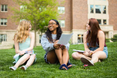 Three young women sit on grass outside a building, holding papers and talking. They appear to be studying, perhaps enjoying a break from their West Chester University housing, with one woman smiling as the others listen attentively.