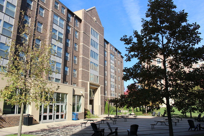A multi-story brick building with large windows, surrounded by trees and an outdoor patio area with tables, chairs, and Adirondack chairs—perfect for relaxing West Chester University housing on a sunny day.