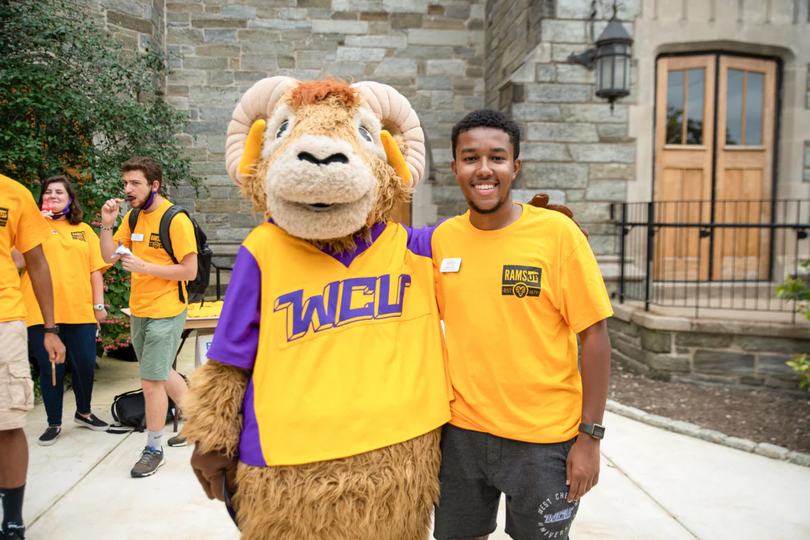 A person in a yellow shirt stands next to a ram mascot in a purple and gold WCU shirt, smiling outside a stone building with wooden doors—an inviting scene near West Chester University housing, with others in yellow shirts nearby.