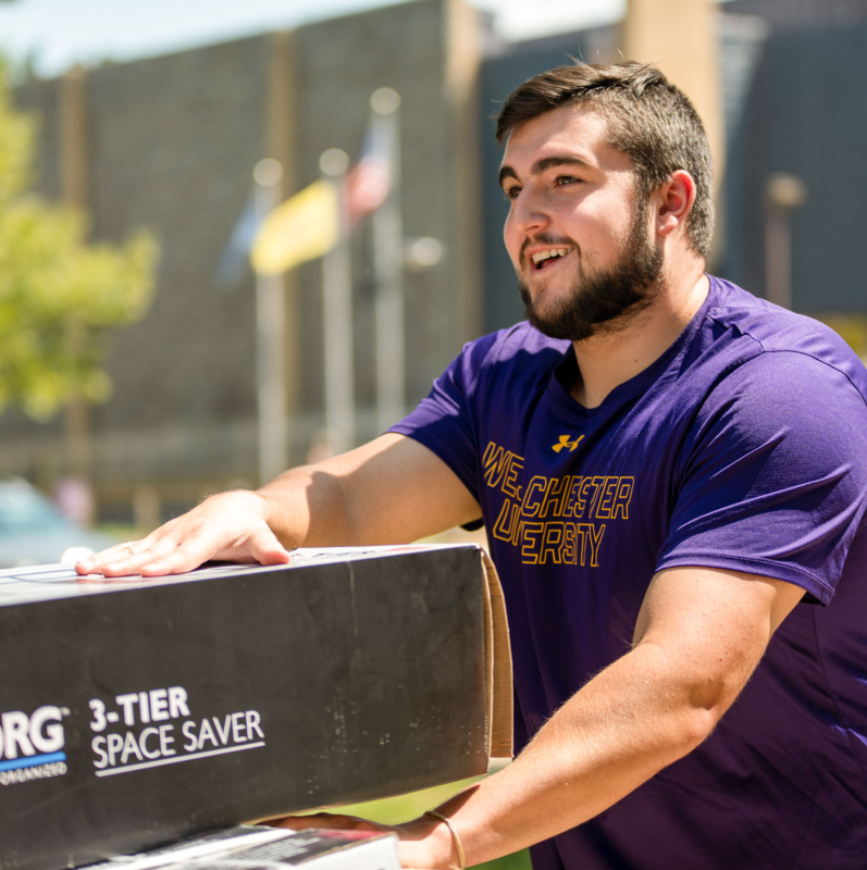 A smiling young man in a purple West Chester University shirt pushes a box labeled 3-Tier Space Saver outdoors on a sunny day, likely moving into West Chester University housing, with flags and buildings visible in the background.