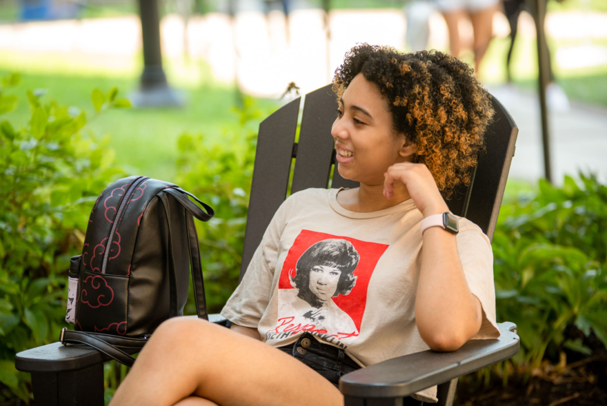 A young person with curly hair sits relaxed in an outdoor chair, smiling and wearing a graphic T-shirt and smartwatch. A black backpack rests on the chair’s arm—a typical scene outside West Chester University housing, with green plants in the background.