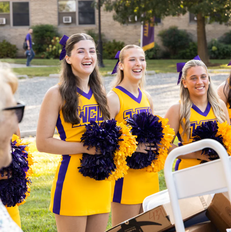 Four smiling cheerleaders in yellow and purple uniforms stand outdoors holding pom-poms, engaging with a woman in the foreground. Trees, a brick building, and West Chester University housing are visible in the background.