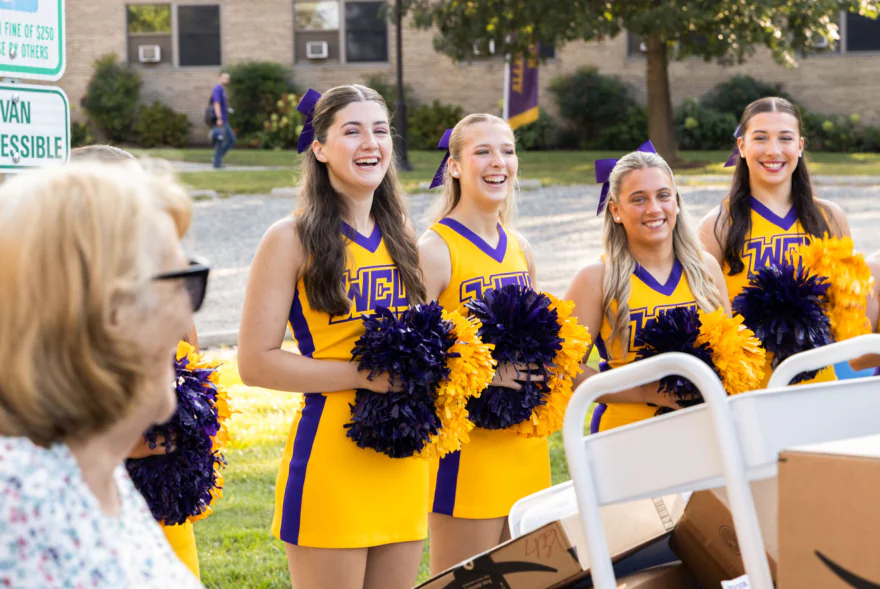 Four smiling cheerleaders in yellow and purple uniforms stand outdoors holding pom-poms, engaging with a woman in the foreground. Trees, a brick building, and West Chester University housing are visible in the background.