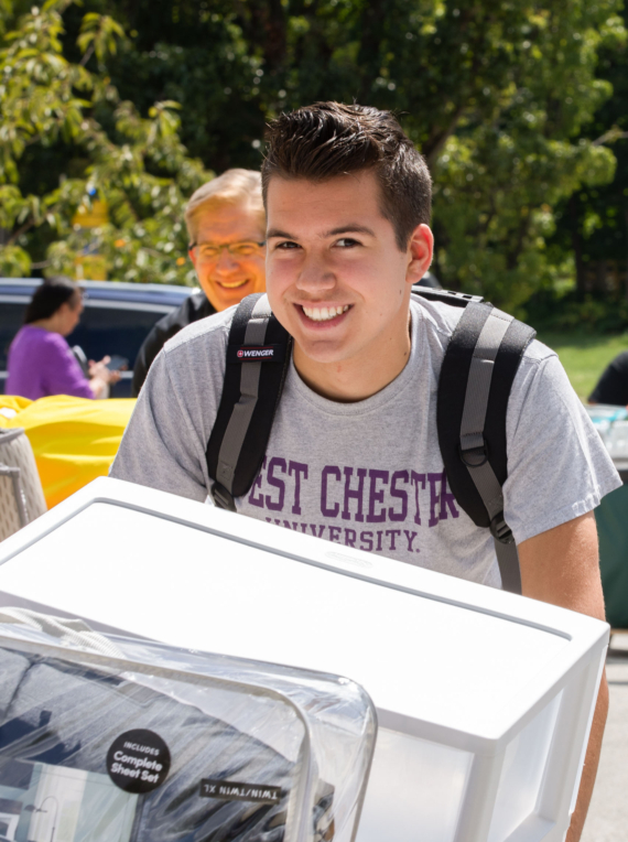 A smiling young man wearing a backpack and a “West Chester University” t-shirt moves a mini fridge outdoors on a sunny day—perhaps moving into West Chester University housing; an older man stands in the background amid trees and cars.