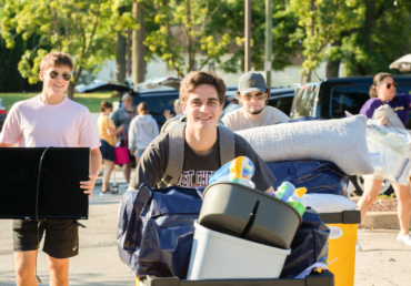 A group of young adults moves belongings outdoors on a sunny day, likely arriving at West Chester University housing. The smiling person in front pushes a cart filled with bedding and storage bins, while others carry items nearby amid trees and parked cars.