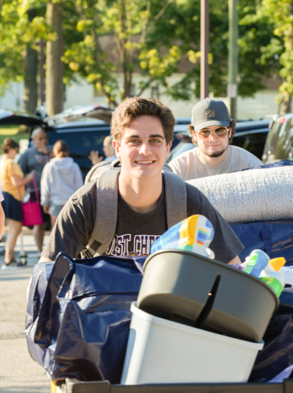 A group of young adults moves belongings outdoors on a sunny day, likely arriving at West Chester University housing. The smiling person in front pushes a cart filled with bedding and storage bins, while others carry items nearby amid trees and parked cars.