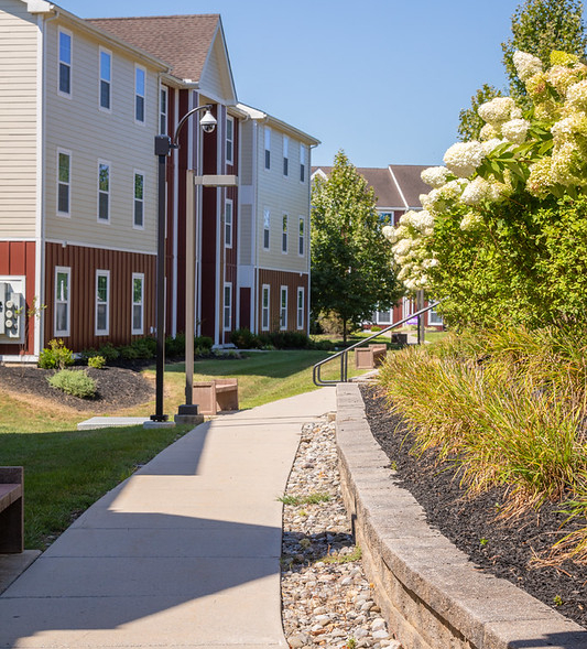 A paved sidewalk curves alongside a low stone wall and landscaped greenery, with multi-story cream and brown apartment buildings—ideal for West Chester University housing—and trees in the background on a sunny day.