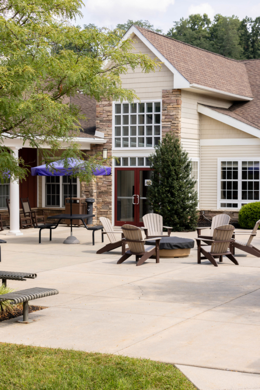 A patio with Adirondack chairs around a covered firepit, benches, and tables with purple umbrellas sits outside a beige building with stone accents and large windows, offering inviting outdoor space for West Chester University housing residents.