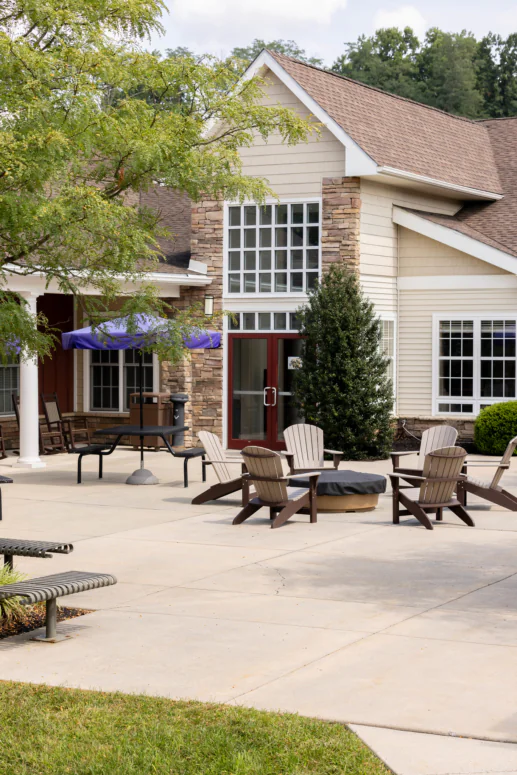 A patio with Adirondack chairs around a covered firepit, benches, and tables with purple umbrellas sits outside a beige building with stone accents and large windows, offering inviting outdoor space for West Chester University housing residents.