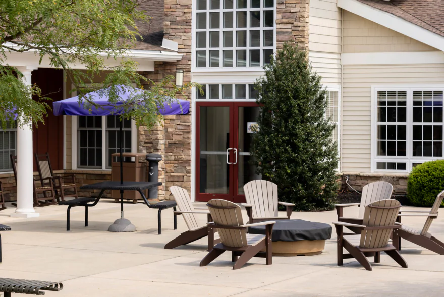 A patio with Adirondack chairs around a covered firepit, benches, and tables with purple umbrellas sits outside a beige building with stone accents and large windows, offering inviting outdoor space for West Chester University housing residents.