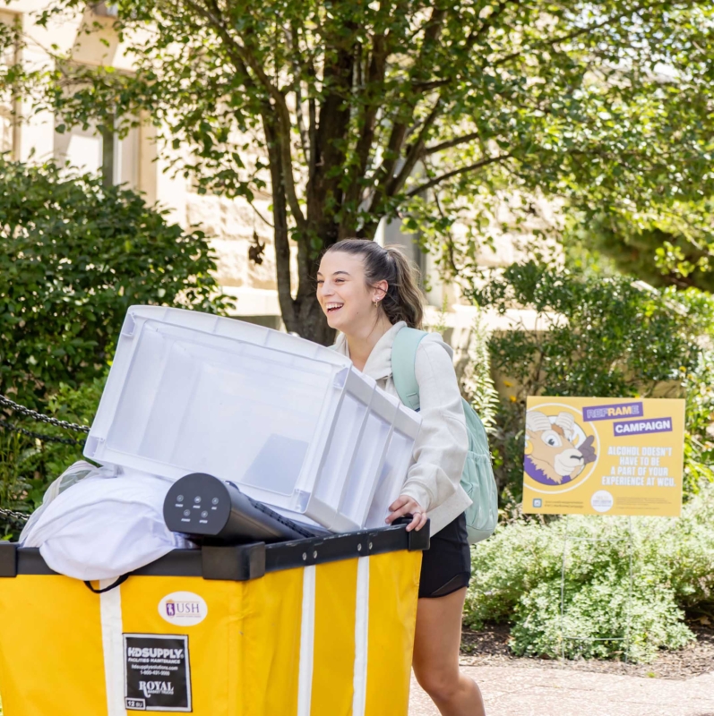 A young woman smiles while pushing a large yellow bin filled with belongings, including a clear storage container and bedding, outside on a sunny day. Trees and a West Chester University housing sign are visible in the background.