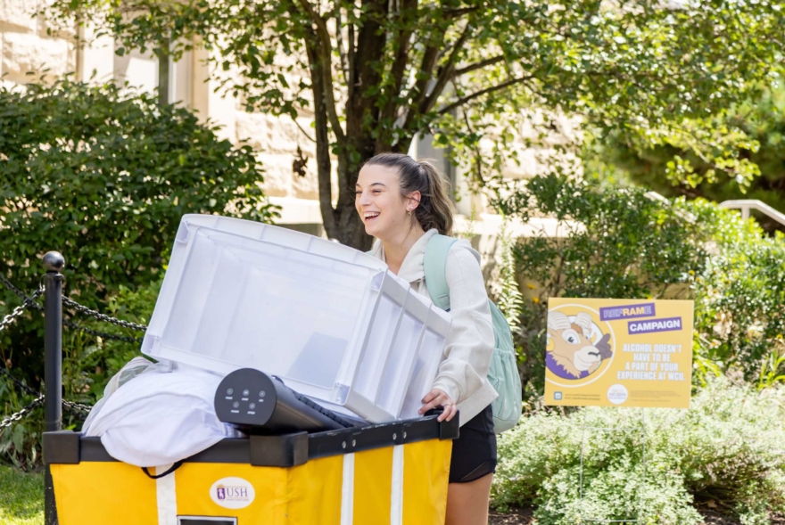 A young woman smiles while pushing a large yellow bin filled with belongings, including a clear storage container and bedding, outside on a sunny day. Trees and a West Chester University housing sign are visible in the background.