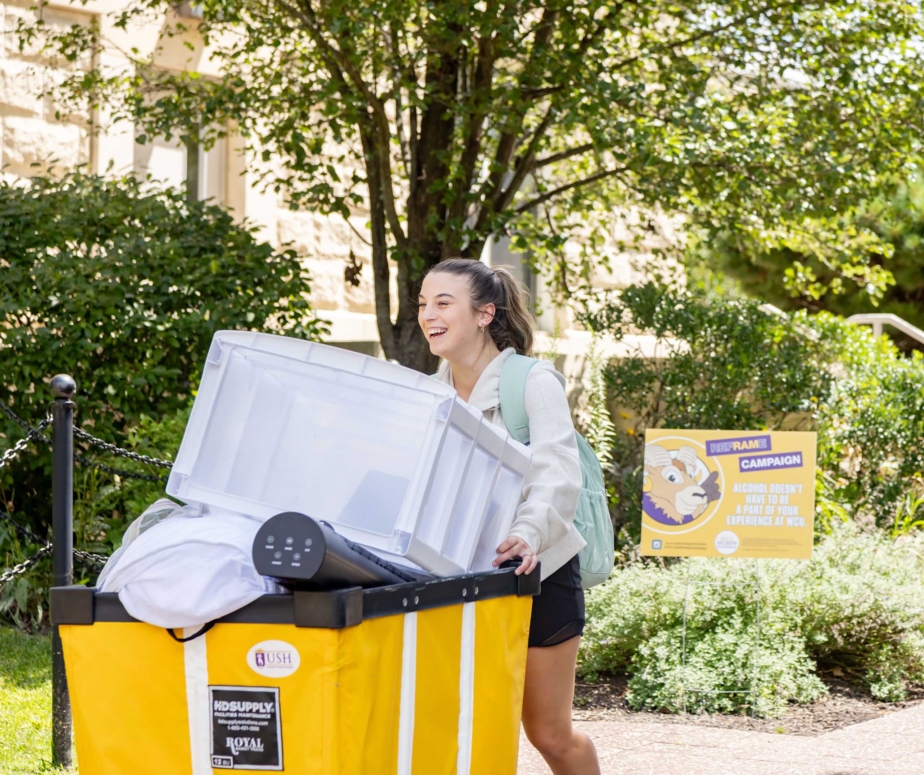 A young woman smiles while pushing a large yellow bin filled with belongings, including a clear storage container and bedding, outside on a sunny day. Trees and a West Chester University housing sign are visible in the background.