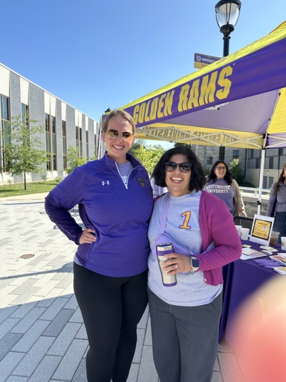 Two women smile and pose in front of a yellow Golden Rams tent outdoors on a sunny day, representing West Chester University housing; one wears a purple pullover, the other a gray shirt with a 1 and purple cardigan, holding a coffee tumbler.