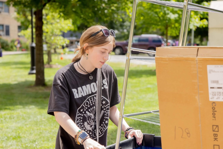 A young person wearing a Ramones T-shirt and sunglasses on their head pushes a cart with a large cardboard box outdoors on a sunny day, moving into West Chester University housing, with green grass and trees in the background.