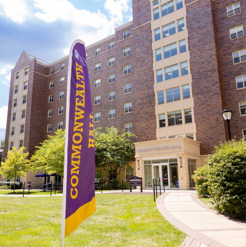 A tall, brick building labeled Commonwealth Hall—part of West Chester University housing—stands behind a purple and yellow flag on a sunny day, with green grass and trees in the foreground.