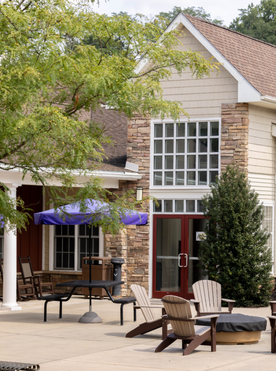 A spacious patio with Adirondack chairs around a fire pit, picnic tables with closed umbrellas, and a beige building with large windows—this inviting outdoor space is perfect for relaxing at West Chester University housing. Green trees and shrubs frame the scene.