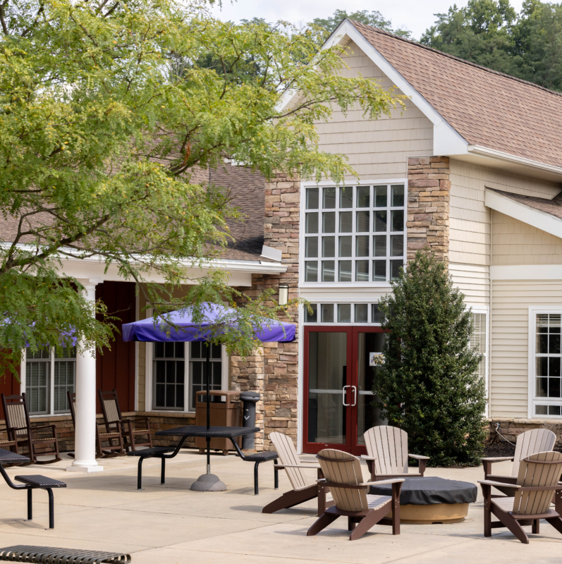 A spacious patio with Adirondack chairs around a fire pit, picnic tables with closed umbrellas, and a beige building with large windows—this inviting outdoor space is perfect for relaxing at West Chester University housing. Green trees and shrubs frame the scene.