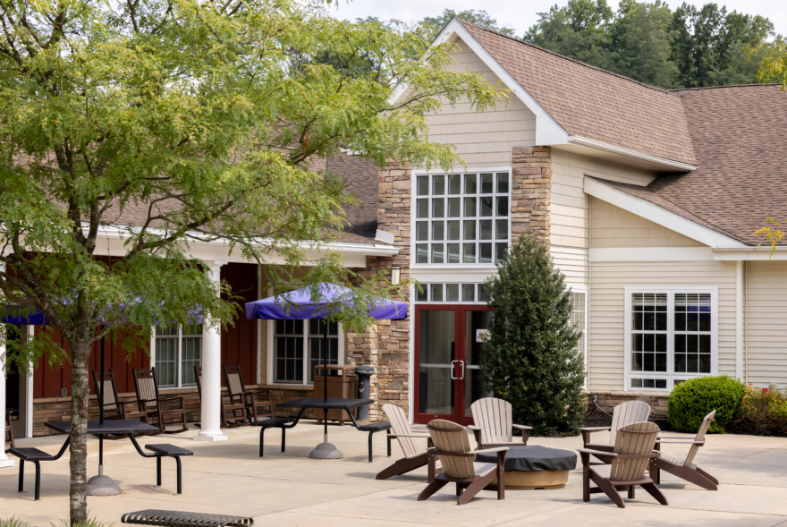 A spacious patio with Adirondack chairs around a fire pit, picnic tables with closed umbrellas, and a beige building with large windows—this inviting outdoor space is perfect for relaxing at West Chester University housing. Green trees and shrubs frame the scene.