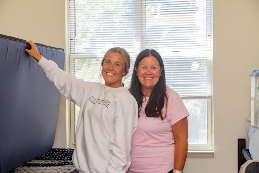 Two women smile in a brightly lit room with a large window behind them; one holds a mattress propped against the wall, suggesting they're settling into their new West Chester University housing.