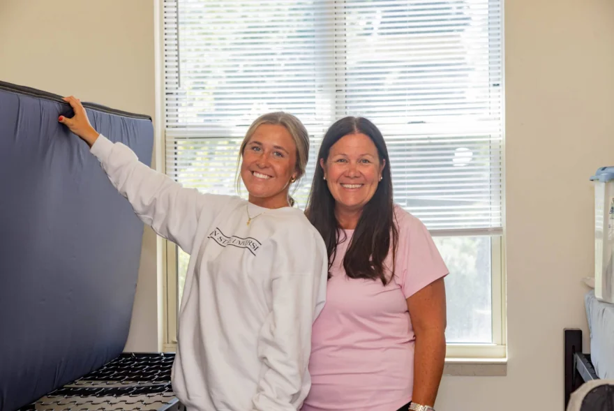 Two women smile in a brightly lit room with a large window behind them; one holds a mattress propped against the wall, suggesting they're settling into their new West Chester University housing.