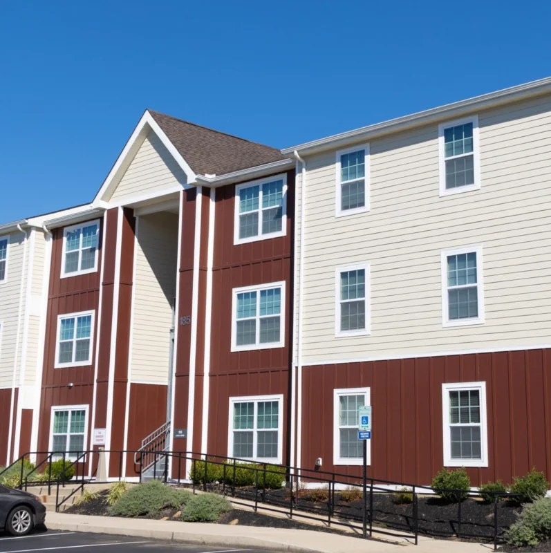 A three-story apartment building with beige and dark red siding, several windows, a ramp for accessibility, and a parked car in front—ideal for West Chester University housing—stands under a clear blue sky.
