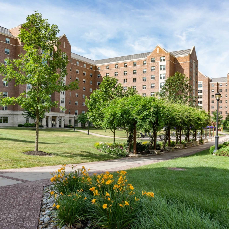 A landscaped college campus courtyard with green lawns, yellow flowers, trees, and brick dormitory buildings under a blue sky with scattered clouds showcases inviting West Chester University housing along a paved walkway.