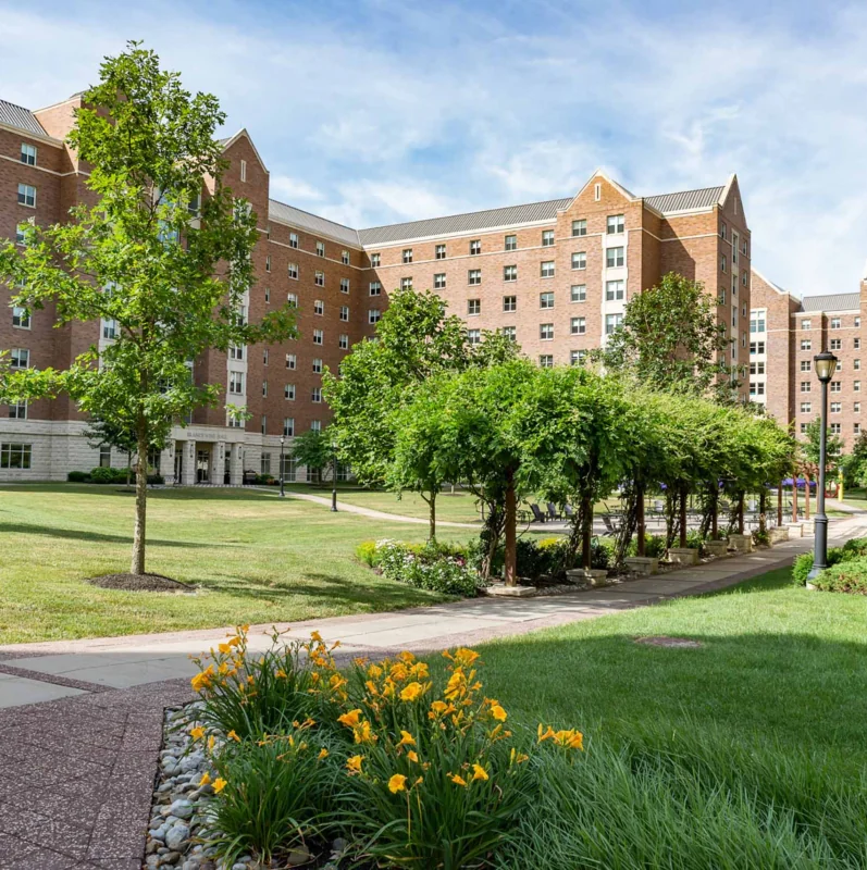 A landscaped college campus courtyard with green lawns, yellow flowers, trees, and brick dormitory buildings under a blue sky with scattered clouds showcases inviting West Chester University housing along a paved walkway.