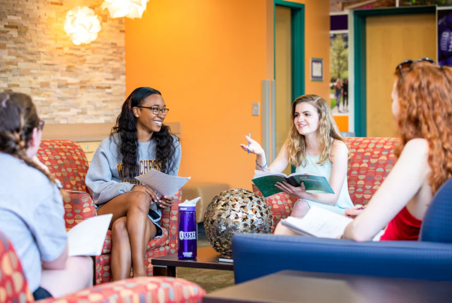 Four young women sit on patterned couches in a brightly lit lounge, talking and holding notebooks. They appear to be engaged in a West Chester University housing study session or group discussion.