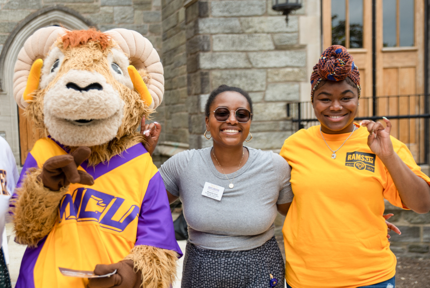 Three people stand smiling outside: a person in a ram mascot costume wearing purple and gold, a person in sunglasses and a grey shirt, and someone in a yellow RAMS UP shirt making a hand gesture—capturing West Chester University housing spirit.