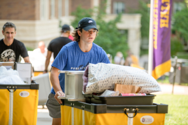 A young person in a blue T-shirt and cap pushes a yellow cart filled with bedding and boxes outdoors on a sunny day, moving into West Chester University housing with others and campus buildings visible in the background.