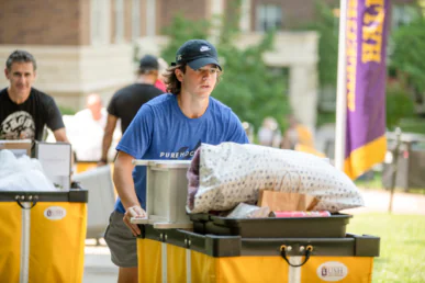 A young person in a blue T-shirt and cap pushes a yellow cart filled with bedding and boxes outdoors on a sunny day, moving into West Chester University housing with others and campus buildings visible in the background.