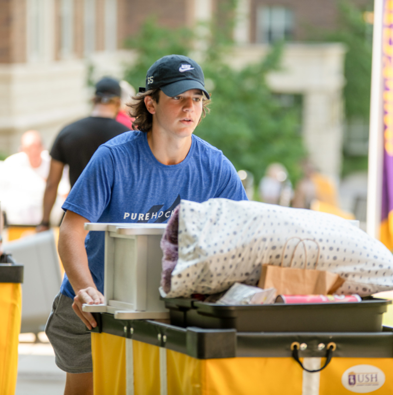 A young person in a blue T-shirt and cap pushes a yellow cart filled with bedding and boxes outdoors on a sunny day, moving into West Chester University housing with others and campus buildings visible in the background.