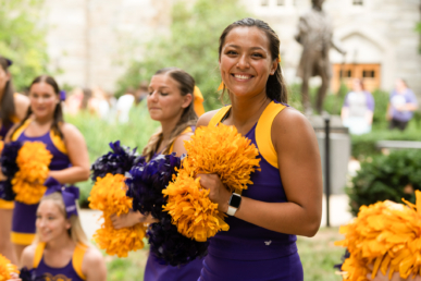 A smiling cheerleader in a purple and yellow uniform holds yellow pom-poms, standing outdoors with other cheerleaders blurred in the background, capturing the spirited energy found near West Chester University housing.