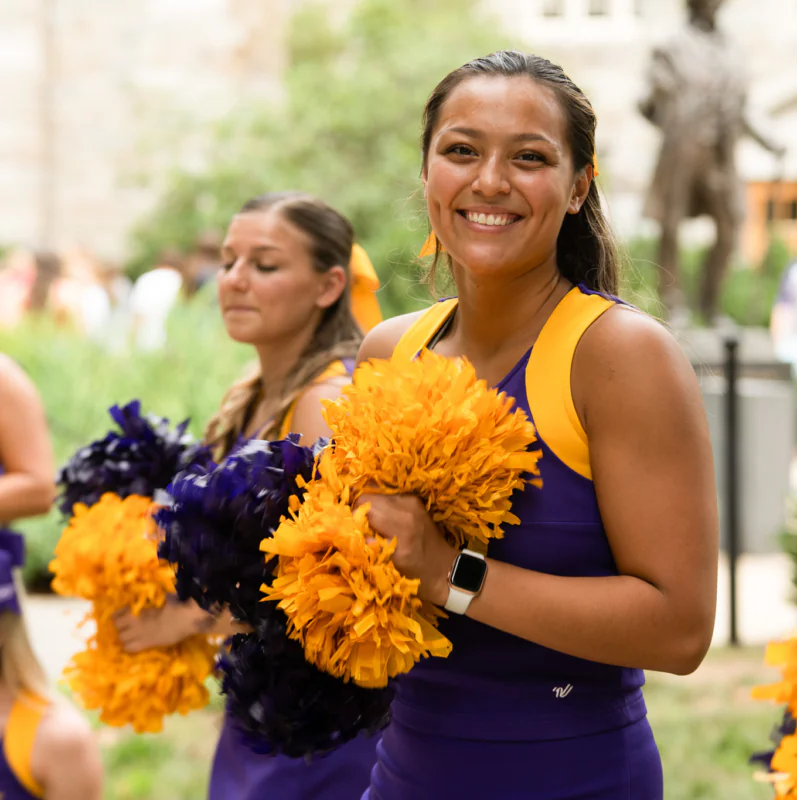 A smiling cheerleader in a purple and yellow uniform holds yellow pom-poms, standing outdoors with other cheerleaders blurred in the background, capturing the spirited energy found near West Chester University housing.