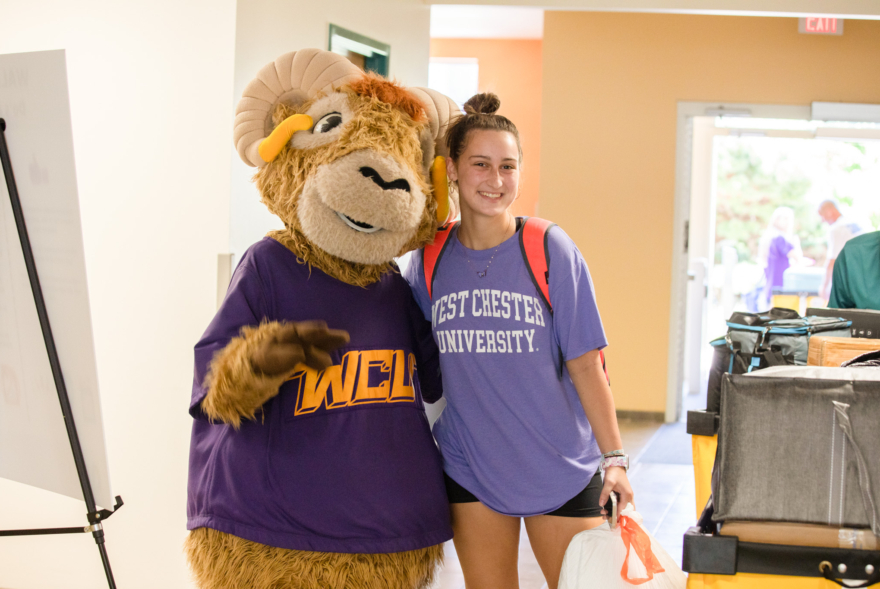A person wearing a West Chester University shirt stands smiling next to a costumed ram mascot inside a brightly lit building, surrounded by luggage and a whiteboard—capturing the excitement of move-in day at West Chester University housing.