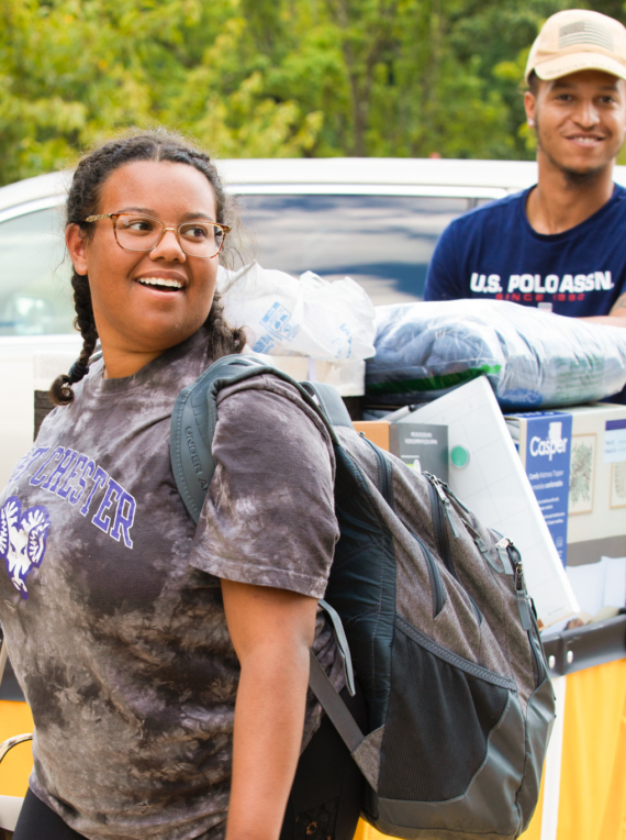 Two young people unload belongings from a yellow cart near a white SUV, moving into West Chester University housing. One smiles while carrying a backpack, and the other stands behind the cart filled with bedding and boxes amid trees and greenery.