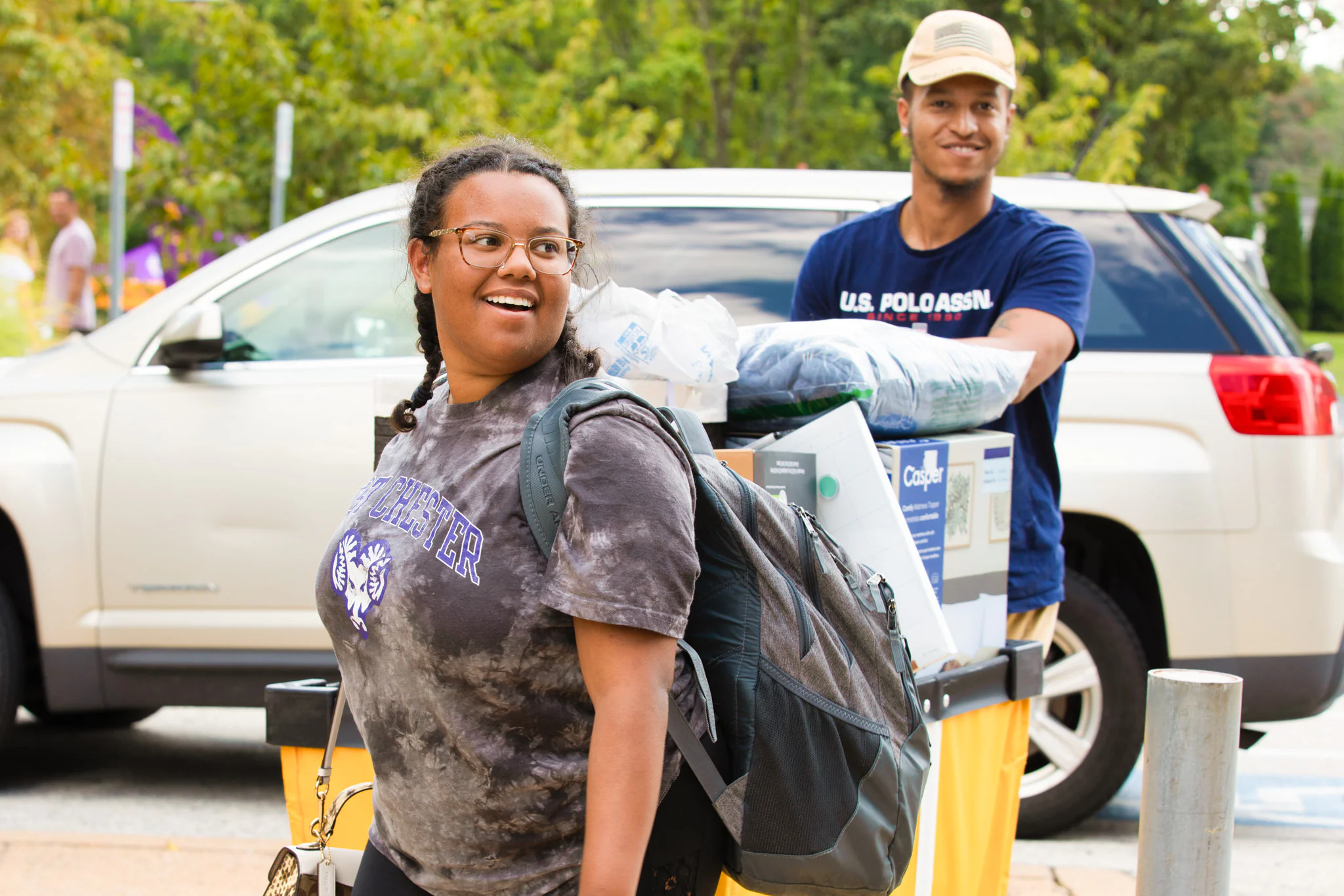Two young people unload belongings from a yellow cart near a white SUV, moving into West Chester University housing. One smiles while carrying a backpack, and the other stands behind the cart filled with bedding and boxes amid trees and greenery.