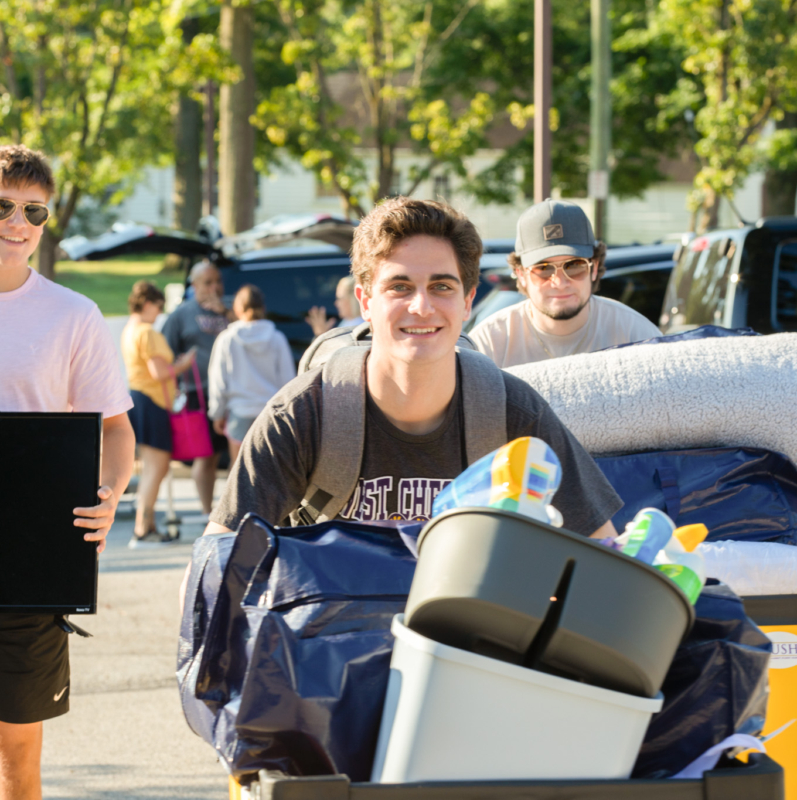Three young men move belongings, including bedding, a laundry basket, and a TV, on a sunny day. Other people in the background also carry items, capturing the lively atmosphere of West Chester University housing move-in day. Trees and cars are visible.